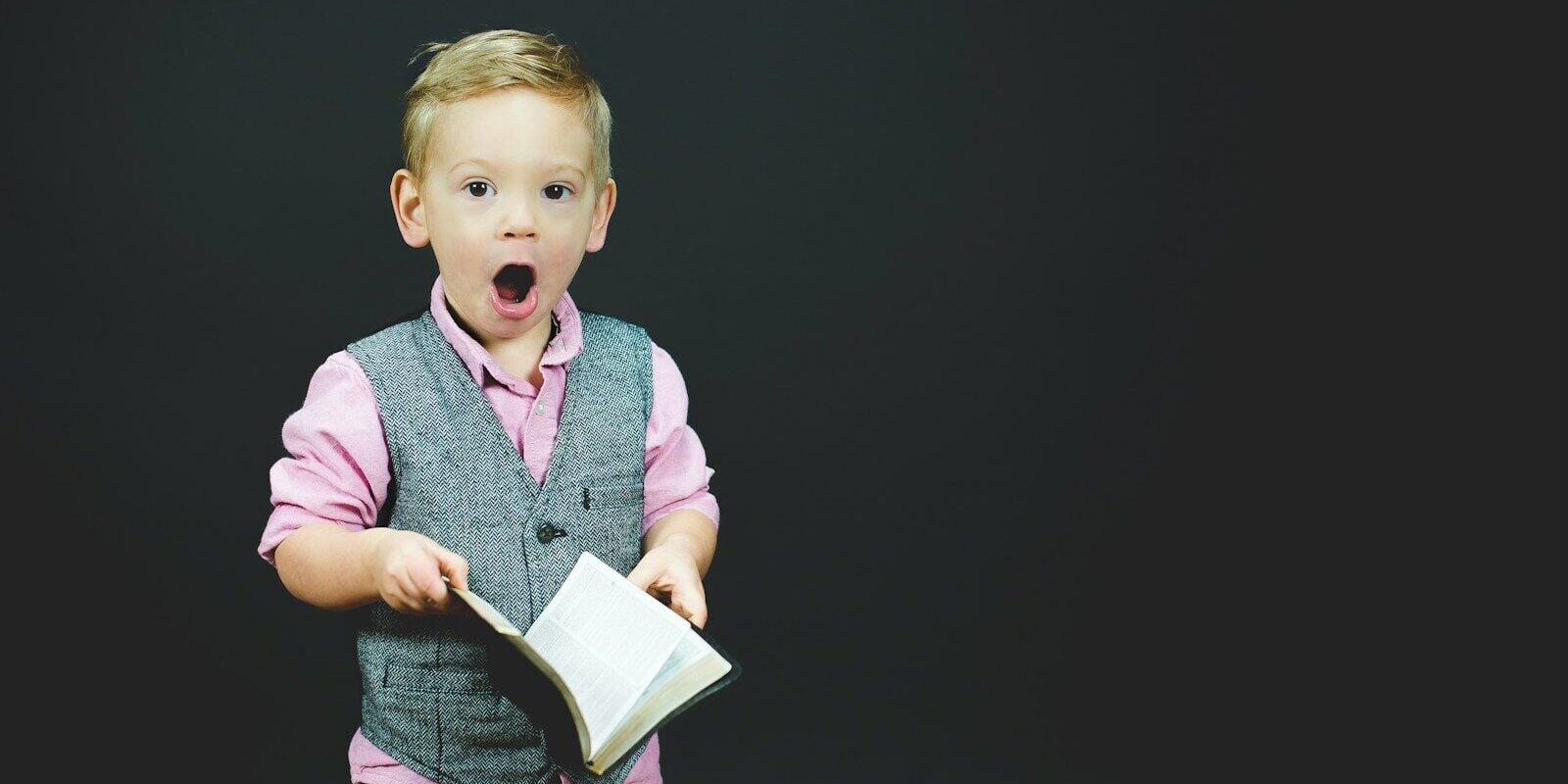 boy wearing gray vest and pink dress shirt holding book